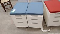 Two white metal filing cabinets with blue cushioned tops side by side on concrete floor in a warehouse setting. Both have three drawers each with metal handles. One cabinet shows some staining on the cushion.