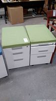 Two white metal filing cabinets side by side, each with three drawers and green cushions on top, floor showing concrete warehouse flooring.