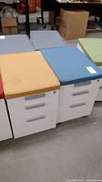 Two metal filing cabinets with cushioned tops, one light brown and one blue, placed side-by-side showing overall condition and key presence.