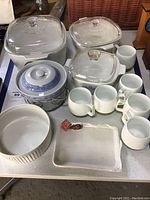 Overview photo showing three CorningWare casserole dishes with glass lids, six white plastic teacups, a blue and white lidded ceramic dish, and a white ribbed ceramic bowl, plus a rectangular ceramic cheese plate.