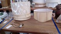 Two metal lamp harps and four different sized and shaped cream colored fabric lamp shades on a wooden surface.