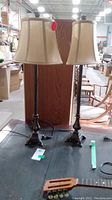 Full view of two metal lamps with beige fabric shades on a black surface, showing overall look and size.