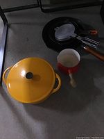 Photo of a yellow Cousances cooking pot with lid, a red metal small pot with white interior and wooden handle, and a vintage round pancake maker with folding handles on the floor.