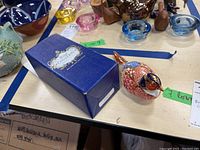 Photo showing the Royal Crown Derby blue box with gold and white emblem and paperweight on the table alongside other background items.