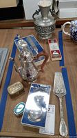 Overview of lot items including silver coffee pot, pewter vase, metal boxes, wine tester and pie server on table with blue painter's tape outlines.