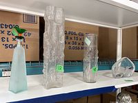 Shelf view of the frosted glass bottle with colorful bird stopper, two textured glass vases (one tall and rectangular, one shorter and cylindrical), and an angular glass piece.