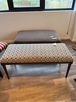 Both cushioned wood bench and cushioned ottoman shown near window on hardwood floor.