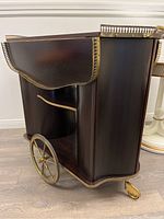 Side view of the wooden bar cart showing drop leaf side in the folded position, brass railing along perimeter, large brass wheels and caster wheel below.