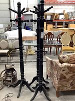 Three black wooden coat racks standing upright in warehouse environment with various furniture in the background.