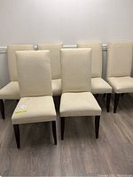 Front view showing six cream cushioned dining chairs arranged in two rows on a wood floor against a white wall.