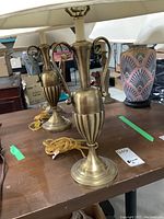 Pair of metal urn-style table lamps on wooden table, showing their design and attached cords.