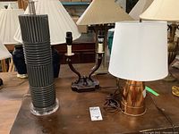 Photo showing three table lamps including a copper pitcher lamp with white shade, a ceramic base lamp, and a metal two-light lamp.
