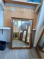 Full view of the large framed mirror leaning against a wooden wall with a wood floor beneath. Decorative frame with gold and bronze carved details visible.