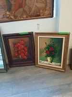 Photo showing two framed floral paintings leaning against a wall on laminate flooring, with size tags on the frames.