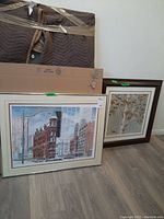 Photo showing two framed prints leaning against the wall on a floor. One depicts the Flat Iron Building and the other a tree.
