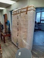Image showing queen size mattress standing upright wrapped in clear plastic secured with brown tape, next to furniture in a room.