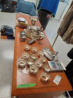 Photo of silverplate teapot, sugar bowl, candy bowl, tray, small pots, butter dish, candlestick, 12 teacups, napkins, and silver care cloth arranged on a wood table.