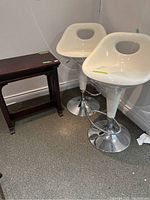 Two white adjustable bar stools and a dark wooden side table with hinged top, arranged in a corner.