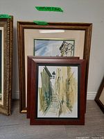 Two framed street landscape paintings, one watercolor with glass and gold-tone frame behind, second is another framed street scene in muted colors, closer front view.