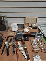 Photo showing seven wristwatches, two table clocks, one small radio, a digital travel clock, eyeglasses, and a compact case.