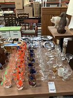 Full view of multiple sets of vintage glassware arranged on a table. Includes tall glasses with orange leaves, shorter glasses with blue grape motifs, clear goblets with gold rims, and various other stemmed glasses.
