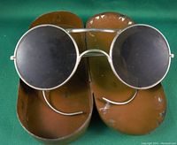 Top view of antique motorcycle goggles showing round dark lenses and brown leather side shields, inside brass case lid visible.