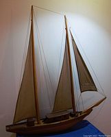 Full model sailboat showing two masts with sails, rigging lines, and wooden hull with cabin details against a plain wall.