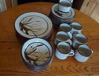 Photo showing stacked dinner plates, lunch plates, cups, and saucers on wooden table with visible brown botanical designs.