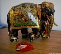 Front and side view of the black lacquered wood elephant figurine with colorful painted scenes and separate broken ear piece.