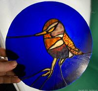 Photo of stained glass round window hanging held up to light showing the colorful hummingbird and blue background.