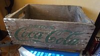 Front view of vintage wooden Coca-Cola crate showing green painted logo on the side with visible aging and wear.