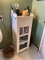 White wooden bathroom cabinet positioned next to a white pedestal sink with a green wall background. Shows three glass panels on the door and a drawer above it.