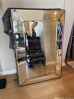 Front view of large rectangular beveled mirror standing upright, reflecting room interior including furniture and artwork.