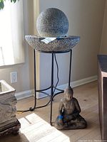 Full view of water feature on black metal stand with seated Buddha statue next to it on floor.