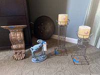 Two metal candle holders with yellow seashell pillar candles, blue metal bird statue, small carved wooden shelf, and round embossed metal plate arranged on carpeted floor.
