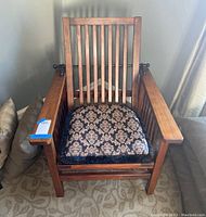 Front view of wooden craftsman style chair with vertical slat back and armrests. Upholstered cushion on seat.