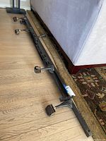 Black metal queen bed frame side rail with attached caster wheels lying on wooden floor next to a couch.