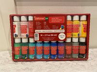 16 bottles of Craft Smart acrylic paint in original packaging, unopened, organized in two rows of eight bottles each showing variety of colors.