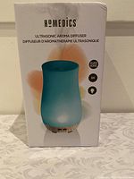 Front view of the boxed Homedics Ultrasonic Aroma Diffuser showing blue handblown glass diffuser with mist output and wood base.