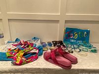 Full view of girls summer items consisting of swimsuit, sandals, multiple sunglasses and goggles, and a beach bag.