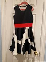 Full view of the girls twirly dress hanging on a pink hanger, showing black bodice, red waistband, and black and white patterned skirt.