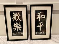 Pair of black framed wall art pieces featuring the words 'HAPPINESS' and 'PEACE' with white Asian characters on a black background and white matting, hung on a white wall.