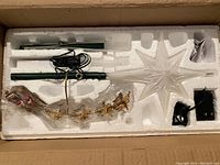 Top-down view of star-shaped tree topper in its original styrofoam packaging with green metal mounting rods, wiring, and certificate.