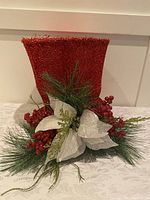 Front view of the red glitter tall top hat adorned with white poinsettias, red berries, and greenery on a white tablecloth background.