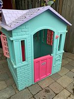 Front view of the LOL Surprise plastic playhouse showing teal walls, pink half door, pink window shutters, and light purple shingled roof.