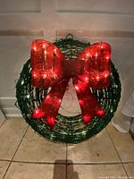 Front view of green wreath with illuminated white lights and large red bow attached.