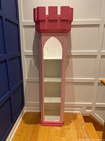 Front view of tall pink and white wooden bookshelf shaped like a tower with an arched opening showing three interior shelves.