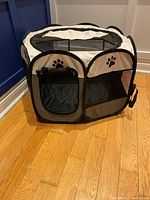 Front and top view of the beige and black small dog foldable playpen showing two zippered doors with paw prints and mesh sides.