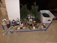 Wide view photo showing assortment of Christmas decorations, lights, and ornaments arranged on floor with bin of string lights