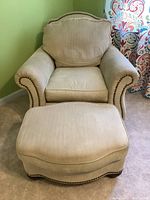 Front view of tan upholstered chair with loose cushions, wood bun feet and nailhead trim alongside matching ottoman.